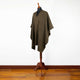 A brown poncho with a subtle pattern is draped over a wooden mannequin stand, standing against a white wall on a polished wooden floor.