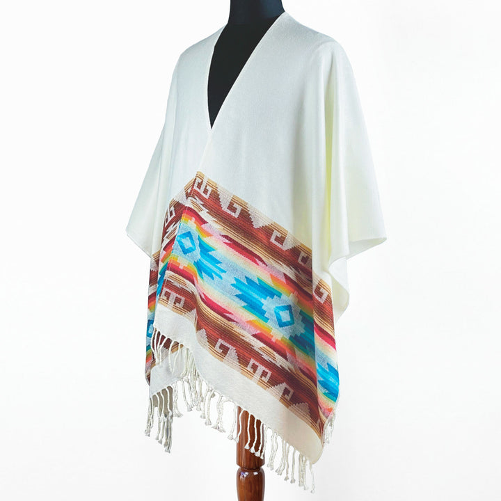 A white poncho features a vibrant, geometric pattern in blue, turquoise, and brown along the lower edge, with fringe detailing. Displayed on a mannequin against a plain background.