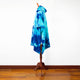 A blue tie-dye poncho with a hood is displayed on a wooden stand, set against a white wall and a hardwood floor.