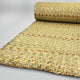 A woven straw mat is partially rolled out, showcasing interlaced patterns of natural fibers. It rests on a neutral, smooth surface, highlighting its texture and craftsmanship.