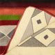 A colorful woven blanket is slightly folded, revealing geometric patterns on the underside. The top side features bold stripes in red, green, and orange hues, while the underside displays diamond shapes.