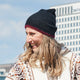 A person wearing a black hat with red trim smiles while outdoors, dressed in a patterned sweater. In the background, a building with several floors is visible.