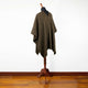 A brown, patterned poncho is draped over a mannequin stand, positioned in a minimalist room with wooden flooring and a plain white wall.