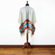 A poncho with geometric patterns and fringes hangs on a wooden mannequin stand, set against a plain white wall on a wooden floor.