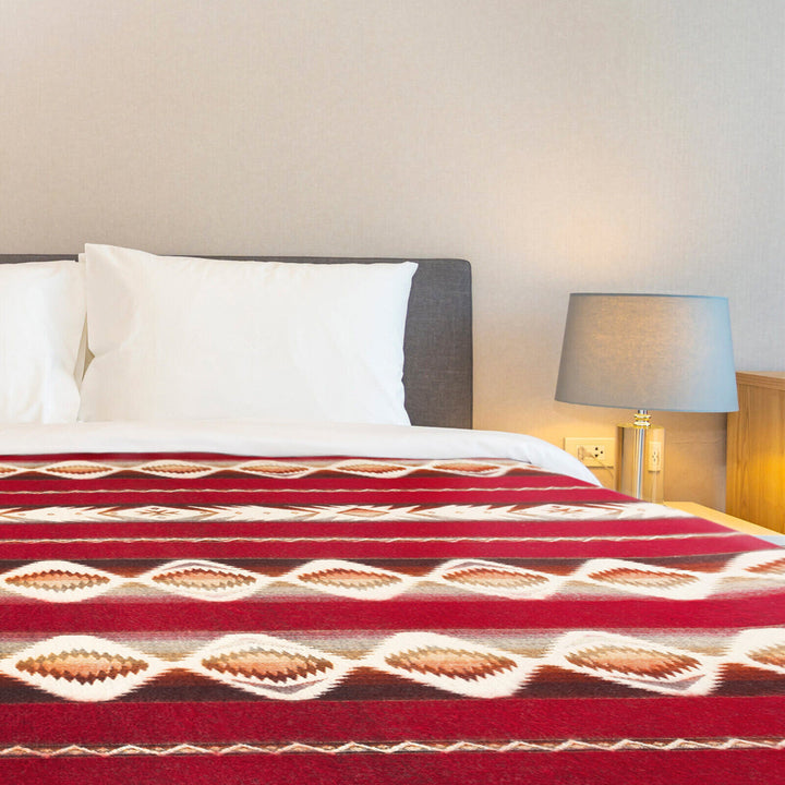 Bed with white pillows and a red patterned blanket, set against a gray headboard in a room with beige walls. A lit lamp sits on a wooden bedside table.