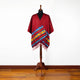 A vibrant red poncho with a colorful geometric pattern and fringe hangs on a mannequin stand against a plain white background, set on a hardwood floor.