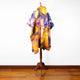 A multicolored, tie-dye cloak with a hood is displayed on a wooden mannequin stand, set against a plain white wall and wooden floor.