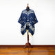 A blue poncho with white geometric patterns hangs on a wooden mannequin stand, set against a plain white wall and wooden floor.
