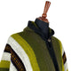 A multicolored woolen poncho with green, white, and dark stripes is displayed on a mannequin. The focus is on the fabric's texture and the poncho's hooded design.