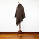 A brown poncho with a hood and fringed edges is displayed on a wooden mannequin stand in a room with a wooden floor and plain white wall.