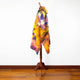 A mannequin displays a vibrant, tie-dye poncho featuring yellow, purple, and orange hues. It's set against a plain white wall, with a wooden floor and tripod stand beneath.