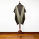 A brown poncho with a geometric pattern is displayed on a wooden stand. It features fringe at the bottom and is set against a white wall and wooden floor.