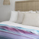 Tinizaray baby alpaca wool blanket – a bed is covered with a striped blanket in shades of blue and purple. Neutral-toned pillows rest against a wicker headboard in a softly lit, white-paneled bedroom