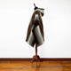A poncho with geometric patterns is draped over a wooden mannequin stand. The background is a white wall, and the floor is wooden.