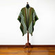 A green poncho with white and orange stripes drapes over a mannequin on a wooden stand, set against a light-colored wall and hardwood floor.