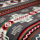 A patterned textile displays multicolored geometric shapes and zigzag lines, predominantly in red, green, and white, on a dark background, creating an intricate and symmetrical design.