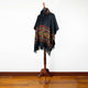A black poncho with colorful geometric patterns draped over a wooden mannequin stand, placed on a wooden floor against a plain white wall.