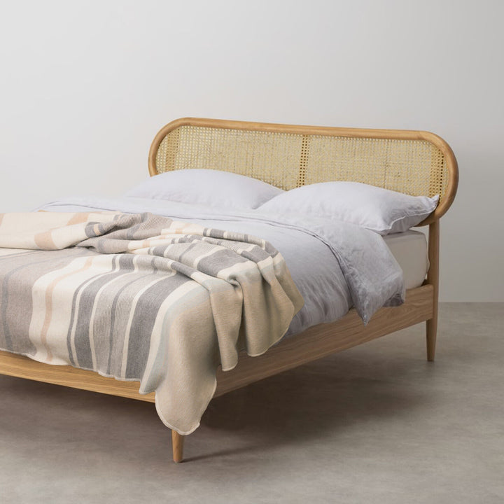 Yankuam baby alpaca wool blanket – a wooden bed with a woven headboard is covered with a striped blanket and white pillows, situated in a minimalist room with a concrete floor and plain white walls