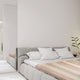 Zhumir baby alpaca wool blanket – a gray upholstered bed with white pillows and beige-striped bedding is positioned in a minimalist, white-walled bedroom. A small side table holds a plant and decorative items