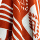 A woolen fabric hangs softly, displaying an intricate pattern of orange and white stripes and geometric shapes, creating a warm, cozy aesthetic against a neutral background.