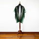 A green and gray poncho with a hood is displayed on a wooden mannequin stand. The setting is a wooden floor with a plain white wall backdrop.