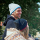 A person wearing a gray hat smiles while sitting outdoors. Another person with long hair, wearing a patterned blanket and black hat, faces away. They are in a park-like setting with trees.