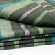 Jambue baby alpaca wool blanket – a stack of folded, woven blankets features stripes in green, blue, and purple hues. The blankets are neatly layered, creating a pattern-rich, textured appearance against a plain white surface