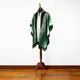 A green and white fringed poncho with a hood is displayed on a wooden mannequin stand in a room with a wooden floor and a white wall background.