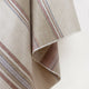 Zhumir baby alpaca wool blanket – a fabric with beige, brown, and gray stripes hangs draped at an angle, with its edges visible, against a plain white background