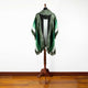 A green and white poncho with geometric patterns, draping over a wooden mannequin stand, is displayed against a plain white wall in a room with a wooden floor.