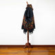 A black poncho with colorful geometric patterns draped over a wooden stand. The poncho's fringed edges hang in a room with wooden flooring and a plain white wall.
