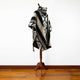 A poncho with geometric black and beige patterns drapes over a wooden mannequin stand, set against a plain white wall and polished wooden floor.