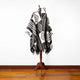 A black and white patterned poncho is draped over a wooden mannequin stand on a polished wooden floor, set against a plain white wall.
