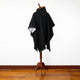 A dark, hooded poncho drapes over a wooden stand, situated in a room with polished wooden flooring and a white wall.