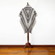 A gray poncho with black and red stripes hangs on a wooden stand, set against a plain white wall and wooden floor.