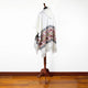 A white poncho with a hood is displayed on a wooden stand, featuring intricate geometric patterns in red, black, and gold. It stands against a white wall on a wooden floor.