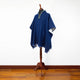 A blue poncho with decorative trim is displayed on a wooden mannequin stand. The setting features a hardwood floor and a plain white wall, providing a minimalistic backdrop.