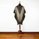 A woven poncho displayed on a wooden mannequin, featuring a geometric, diamond-patterned design in brown and beige on a hardwood floor with a plain white background.