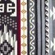 A colorful textile features geometric patterns with bold black, white, red, and gray designs, including stripes, diamonds, and wavy lines. The pattern alternates vertically, creating a dynamic visual effect.