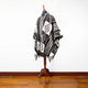 Poncho displayed on a wooden stand, features bold geometric patterns in black and white, with fringed edges. It's situated on a polished wooden floor against a plain white wall.
