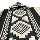 Poncho with a black and white geometric pattern, featuring diamond and zigzag designs. The garment has a hood and wooden toggle buttons, laid flat against a white background.
