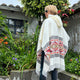 Person wearing a patterned poncho stands outdoors. They are surrounded by lush greenery and flowers next to a building, suggesting a garden or courtyard setting.