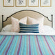 Tzacantza baby alpaca wool blanket – a bed displays a striped blue and purple blanket with decorative pillows, including a blue-striped one, set against a beige upholstered headboard. Framed botanical prints hang above