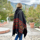A person wearing a patterned black poncho stands facing away, in a cobblestone courtyard surrounded by potted plants and green foliage, with mountains visible under a cloudy sky.