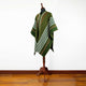 A green poncho with white and brown stripes is displayed on a wooden mannequin stand, set against a white wall and wooden floor background.