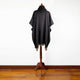 A dark-colored poncho with fringe edges drapes over a wooden mannequin stand, positioned on a polished hardwood floor against a plain white wall background.