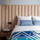 Bed with blue pillows and geometric blanket, placed against a vertically tufted tan headboard. A side table holds a vase with flowers and magazines. Hanging lights are partially visible.
