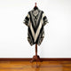 A poncho with geometric patterns drapes over a mannequin stand, displayed in a minimal indoor setting with a wooden floor.