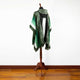 A green and white poncho with fringed edges displays on a wooden stand, set against a white wall and wooden floor.