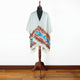 A white poncho with colorful geometric patterns drapes over a wooden mannequin stand, set against a plain white wall and hardwood floor background.